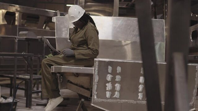 Full shot of female African dockyard worker wearing green overalls, hard hat and safety goggles, sitting on stern of metal framework of boat, writing