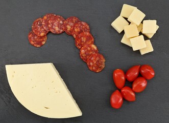 Slices of spicy salami with parmesan cubes, soft cheese and cherry tomatoes on a black slate serving plate