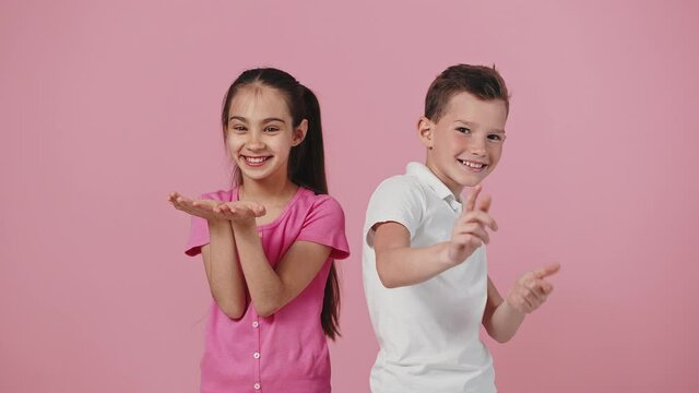 Cute positive brother and sister posing to camera, girl sending blow kiss, boy pointing fingers, pink studio background
