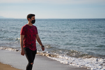 Man walking on the seashoreA man walking along the seashore barefoot. Caucasian man in red T-shirt and black trousers. On a sunny day on the sandy beach. Wearing a watch and mask. Denia.
