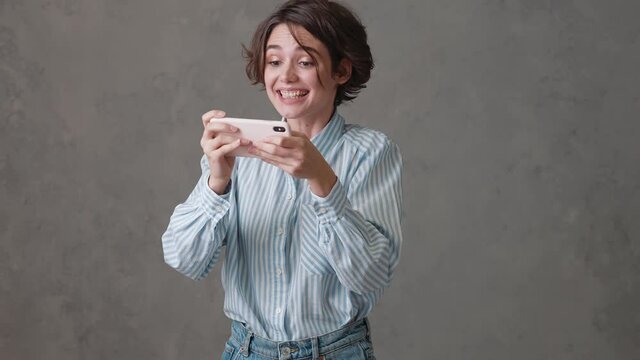The enthusiastic woman playing on the phone and smiling while standing in a gray studio