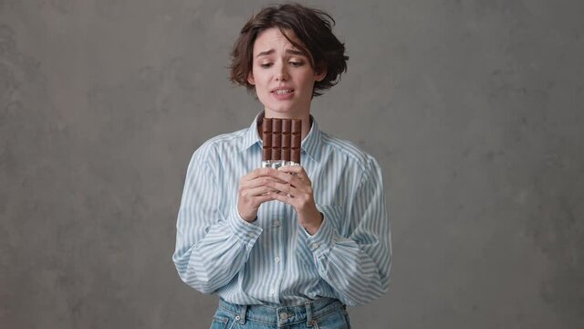 A upset woman with short hair holding a chocolate bar in her hands and wanting to take a bite while looking to the sides in a gray studio