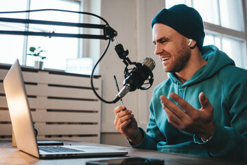 Cheerful host with stubble streaming his audio podcast using microphone and laptop at his small...