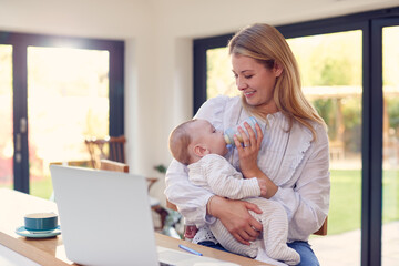 Working Mother Using Laptop At Home Whilst Feeding Baby Son On Knee With Bottle