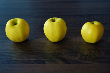Golden delicious apples on a dark table.
Three sweet yellow fruits arranged in a row on a dark painted wooden background. Wallpaper with natural products and fruits.  
