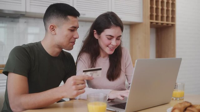 The smiling couple writing down a bank card number on the laptop at the breakfast while sitting at the table in the kitchen