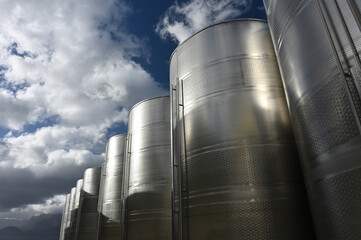 A row of outside stainless steel fermentation wine tanks against  clouds and a blue sky