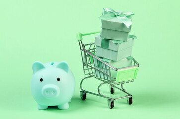 Holiday shopping. Mini supermarket trolley with gift boxes and piggy bank on green pastel background
