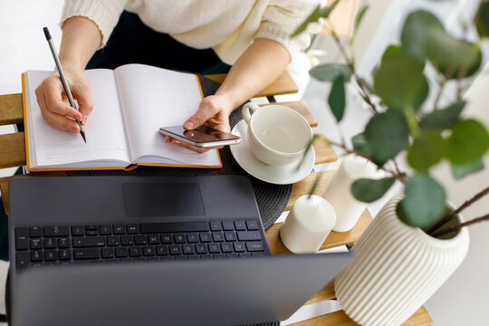 business woman works at laptop, holds smartphone in her hand and makes entries in diary. Business planning and tasks for day or week. Female in business - Powered by Adobe