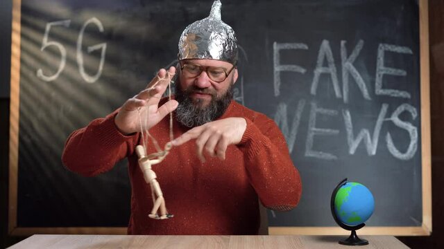 A man video blogger sits at a table against the background of a chalk board with the text Fake News. a conspiracy theorist guy in a foil hat holds a wooden man's puppet on strings in his hands.