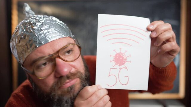 Caucasian male blogger is sitting at home, a hat with foil is worn on his head. Conspiracy theory. Holds a tablet with the text 5g.