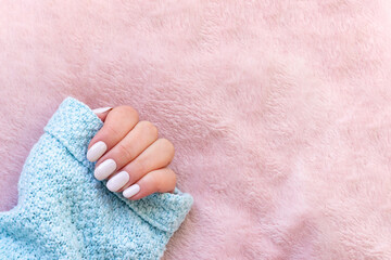 Female hand in blue knitted sweater with beautiful manicure - white ivory nails on pale pink fluffy fabric, textile background with copy space
