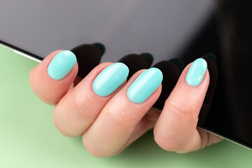 Female hand with beautiful manicure - mint blue nails with smartphone on green background. Selective focus