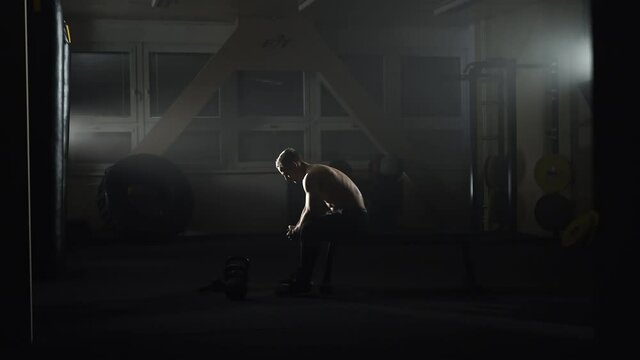Bodybuilder sitting on a gym bench, contemplating in shadows.