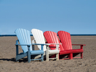 Red, white and blue Adirondack chairs on a sandy beach on a sunny day
