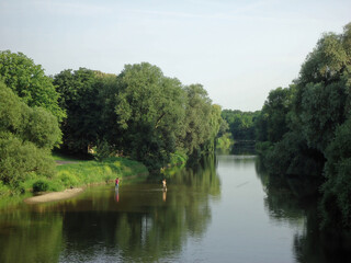 Rzeka w Alzacji, brzegi poro śnięte drzewami, dwie osoby moczą nogi w wodzie.