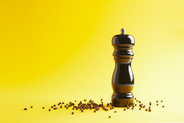 Close up view of black pepper mill with peppercorns on yellow background.