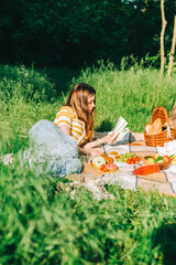 Young woman reading book outdoors, in natural park on picnic, lying on a picnic blanket and...