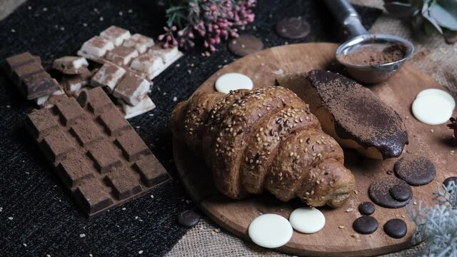 Sweet food composition. Fresh baked Croissant, piece of fresh bread with chocolate and cocoa powder on a wooden board, white and dark chocolate on a table. Delicacy and yummy dessert. 4k footage