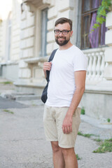Man smiles and looks directly into the camera with a beard in glasses, a white t-shirt, beige shorts