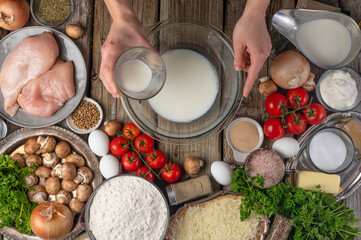 In the photo, the chef prepares the dough. He pours milk into a bowl. Very bright foods are spread around. Wooden table. Many different products. View from above.