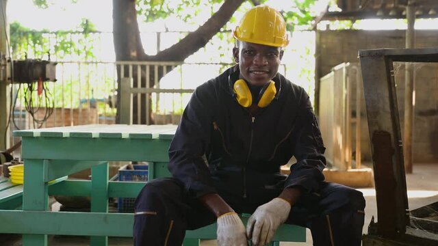 Portrait of African American workman wearing uniform and yellow safety hardhat sitting smiling on table in factory. Happy African young engineer sitting and holding big adjustable wrench in workplace.