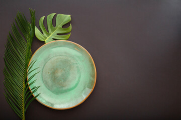 Empty green plate on black background. Luxury dinner plate served with asian kinfolk grunge style tropical palm leaves monstera plant. Mock up empty ceramic plate. Top view copy space