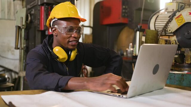 African engineer man in uniform wearing safety helmet, goggle and sitting using computer laptop for talking video conference meeting about production plan in workplace at factory. Technology concept. 