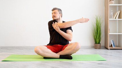 Fototapeta premium Home training. Sportive man. Fitness gym. Lockdown lifestyle. Calm concentrated guy in t-shirt shorts sitting lotus pose doing stretching workout for hands in light room interior.