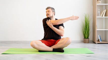 Home training. Sportive man. Fitness gym. Lockdown lifestyle. Calm concentrated guy in t-shirt shorts sitting lotus pose doing stretching workout for hands in light room interior.