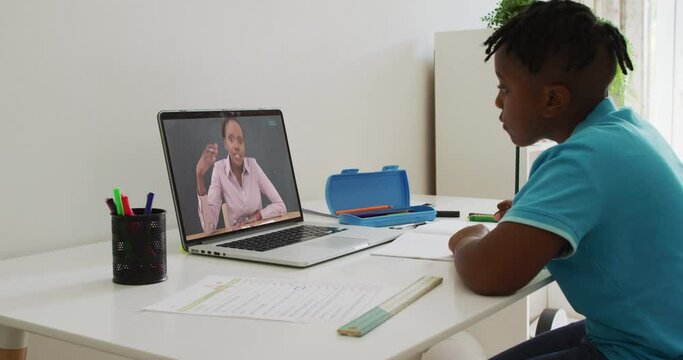 African american boy doing homework while having a video call with female teacher on laptop at home