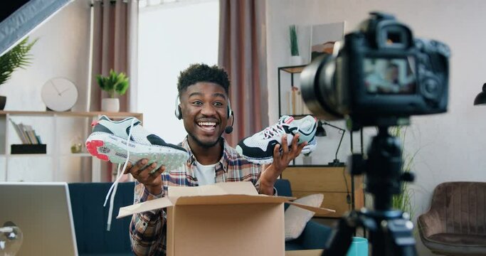 Handsome happy smiling young black-skinned guy in wireless headset recording video for his internet audience while showing his new sneakers from delivery parcel