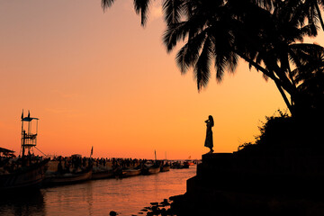 A female and a palmtree silhouettes in front of boats against an orange sunset in GOA