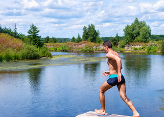 Fototapeta na wymiar Teenager running on a wooden bridge, sprints to jump into the river. Sports guy jumping into the water on a sunny day. Beautiful landscape of a river with green banks. Concept of healthy lifestyle