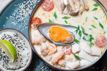 Closeup view of tom yum or tom yam hot, spicy and sour Thai soup with seafood, shrimps (prawn) and mussels decorated with scallions served in bowl with cooked boiled rice garnished with sesame seeds