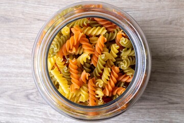 A top down portrait of a glass pot, jar or bowl full of spirelli in three different colors. The uncooked tricolore italian pasta variant is orange, yellow and green and is swirl shaped.