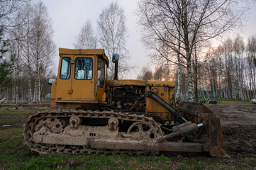 An old rusty yellow tractor among the trees in the open air on a driverless farm. Agricultural machinery