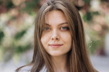 Close up portrait attractive young adult woman model looking at camera, happy beautiful 20s brunette lady pretty face dental smile posing stand alone at park on a background of sakura trees