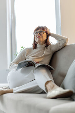 middle-aged adult woman with glasses sits on the sofa and reads a magazine
