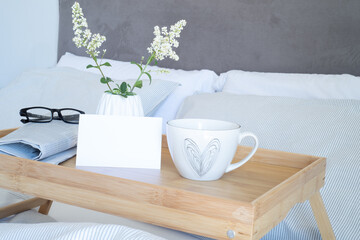 Breakfast in bed stationery mockup scene. Blank greeting card mockup, cup of coffee, newspaper and glasses on tray. White vase with small flowers. Valentine's day breakfast, lifestyle concept.
