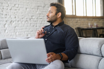 Contemplative mixed-race hindu guy sits on the sofa with a laptop and waiting for inspiration, looks away, figured out how to solve an important problem or project issues