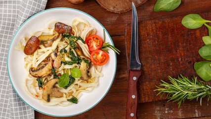 Udon with mushrooms and spinach in a plate on a gray napkin next to a knife and mushrooms and herbs.