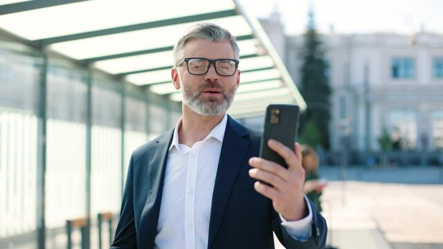 Close up portrait of cheerful Caucasian middle-aged handsome stylish man videochatting on smartphone online standing in town on train station, traveling, businessman speak on video call on cellphone