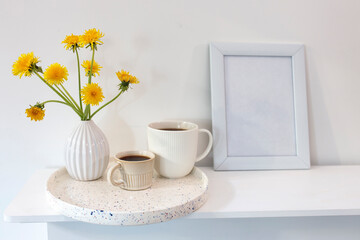 A bouquet of dandelions in a white fluted vase and two cups of different sizes with coffee on a white table.