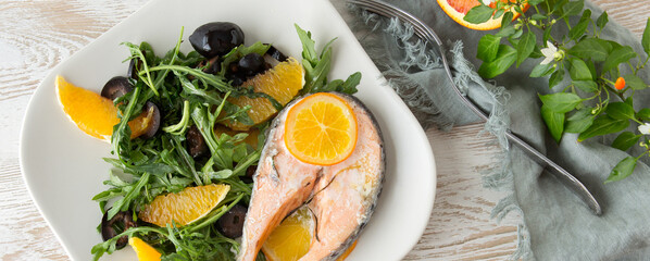 Fototapeta na wymiar flat lay of plate of baked salmon steak with oranges, olives and arugula on light wooden table
