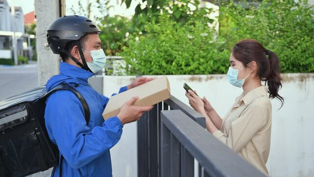 Asian courier man in uniform wear medical face mask and helmet using bicycle with backpack for food delivery of order to customer. Young woman receive food to order from courier on during quarantine. 