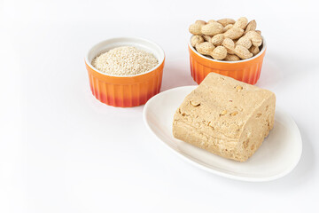 Sesame-peanut halva lies on a white plate on a white background. Next to it are orange ramekins with sesame seeds and peanuts. Traditional national dessert. Oriental, Turkish and Arabic cuisine.