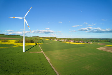 1 wind turbine on green farmland. Small village in the background. Blue sky with small moon. Aerial view. Front view.