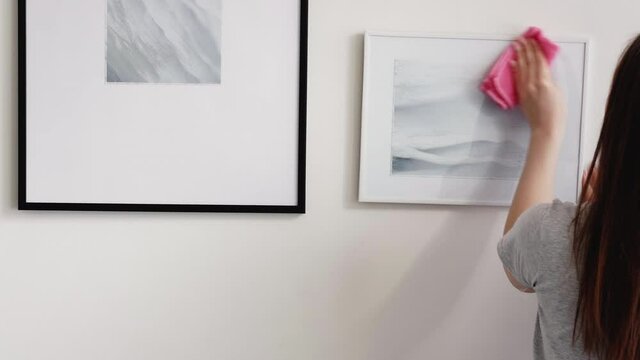 Home cleaning. Female worker. Room service. Perfect interior. Unrecognizable woman in light gray t-shirt removing dust from painting art with microfiber duster patch.