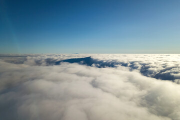 Aerial view from above of white puffy clouds and distant mountain top in bright sunny day.